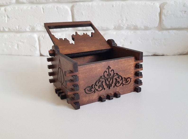 Pu&ograve; includere: Una scatola di legno con un coperchio incernierato, che presenta un design intagliato di un castello sul coperchio e un motivo floreale decorativo sul fondo. La scatola &egrave; di colore marrone e ha una finitura in legno scuro. Il lato della scatola presenta un'immagine intagliata del simbolo dei Doni della Morte.