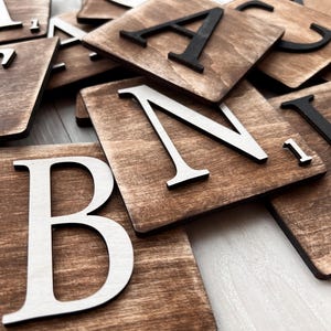 May include: Wooden letter tiles with a dark brown stain and contrasting white or black letters. The letters are in a serif font and are scattered across a light surface. The tiles are square and appear to be for crafts or decor.