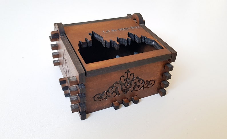 Pu&ograve; includere: Una scatola di legno con un ritaglio del castello di Hogwarts e il testo "I Am The Magic One". La scatola ha un design decorativo sul fondo.