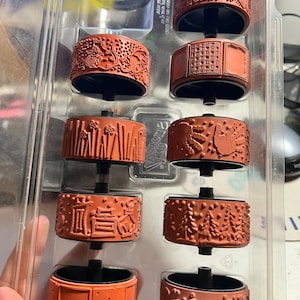 May include: A set of ten red rubber stamps with various designs, including a tree, a handprint, a house, and a grid pattern. The stamps are in a clear plastic storage case.