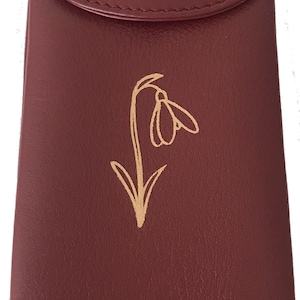 May include: A burgundy leather case with a flap closure. A gold-colored illustration of a snowdrop flower is on the front. The case is rectangular and appears to be for glasses or a similar item.