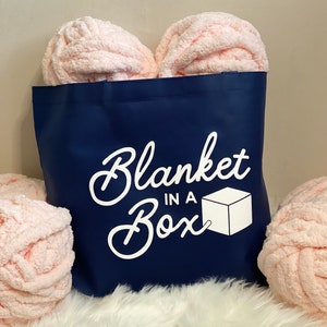May include: A navy blue tote bag with the text "Blanket in a Box" in white lettering. The bag is filled with three pink fluffy yarn balls.