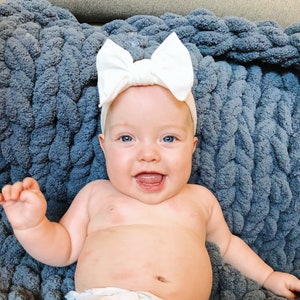 May include: A baby wearing a white bow headband and a diaper, lying on a blue knitted blanket. The baby is smiling and looking at the camera.