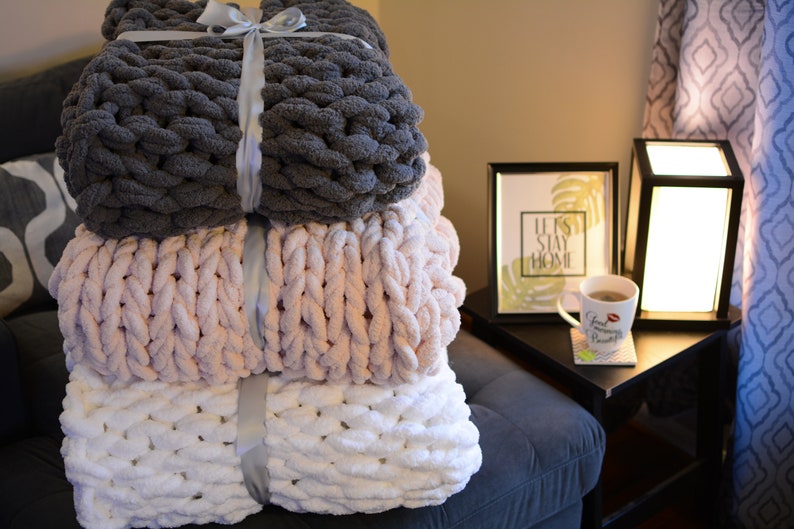 May include: Three chunky knit blankets stacked on a couch. The top blanket is gray, the middle blanket is pink, and the bottom blanket is white. The blankets are tied with a gray ribbon.