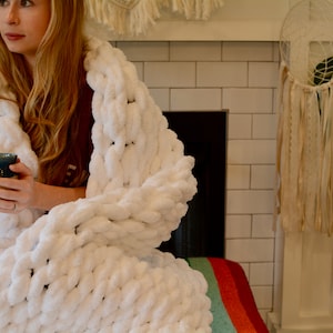 May include: A white chunky knit blanket draped over a person holding a blue and white ceramic mug. The blanket is made with large, thick stitches and has a soft, fluffy texture.