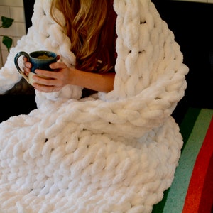 May include: A white chunky knit blanket draped over a person sitting on a colorful striped bench. The person is holding a blue and white ceramic mug.