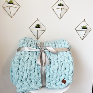 May include: A light blue chunky knit throw blanket tied with a silver ribbon. The blanket is sitting on a white stool with wooden legs. There are three geometric wall planters with plants in them.