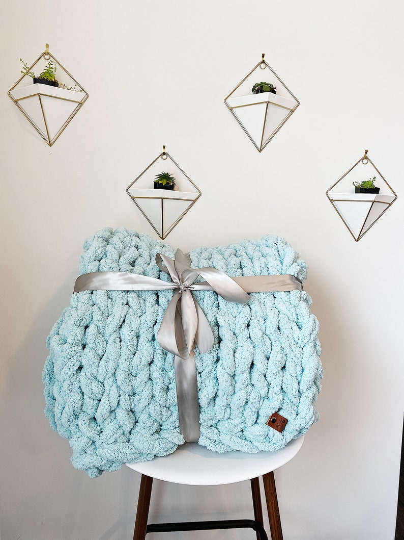 May include: A light blue chunky knit throw blanket tied with a silver ribbon. The blanket is sitting on a white stool with wooden legs. There are three geometric wall planters with plants in them.