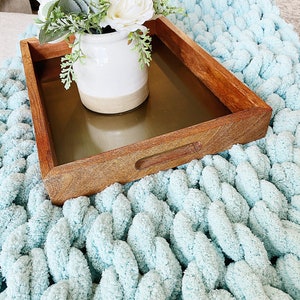 May include: A wooden tray with a gold metal insert, featuring a white ceramic vase with a bouquet of white flowers and green leaves. The tray is sitting on a light blue chunky knit blanket.