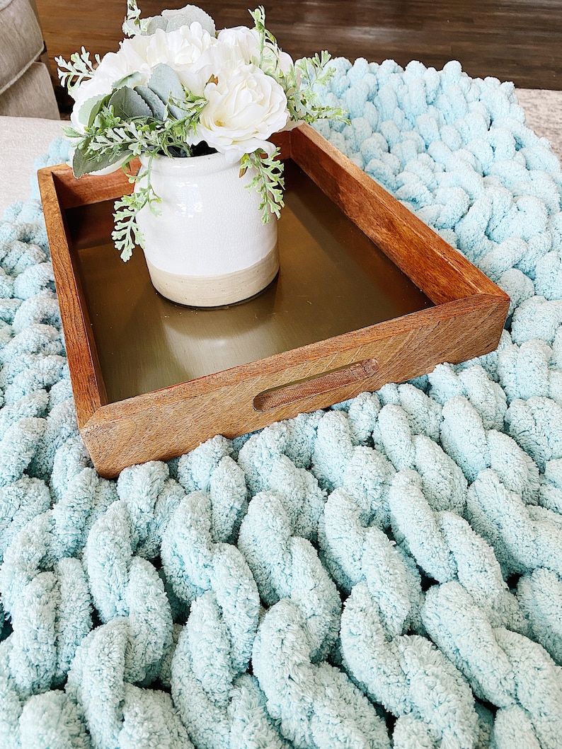 May include: A wooden tray with a gold metal insert, featuring a white ceramic vase with a bouquet of white flowers and green leaves. The tray is sitting on a light blue chunky knit blanket.