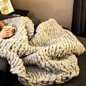 May include: A person is sitting on a couch with a gray chunky knit blanket. The blanket is soft and fluffy, and it is draped over the person's legs. The person is holding a mug in their hands.