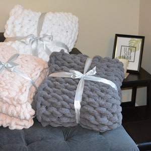 May include: Three chunky knit throw pillows in white, pink, and gray are tied with white satin ribbon. The pillows are arranged on a gray couch with a black end table and a framed print that says "Let's Stay Home".