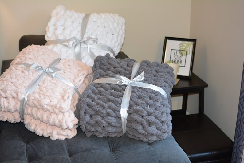May include: Three chunky knit throw pillows in white, pink, and gray are tied with white satin ribbon. The pillows are arranged on a gray couch with a black end table and a framed print that says "Let's Stay Home".