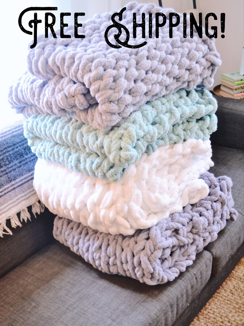 May include: A stack of four chunky knit blankets in various colors, including gray, light blue, white, and purple. The blankets are arranged on a gray couch. The text "FREE SHIPPING!" is displayed above the blankets.