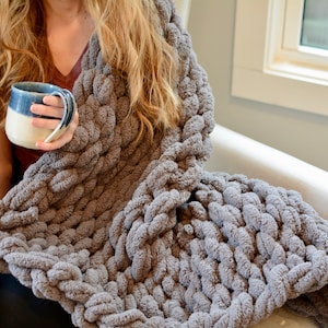 May include: A person is sitting in a chair wrapped in a gray chunky knit blanket. The blanket is made of thick, soft yarn and has a textured, cable knit pattern. The person is holding a ceramic mug with a blue and white glaze.