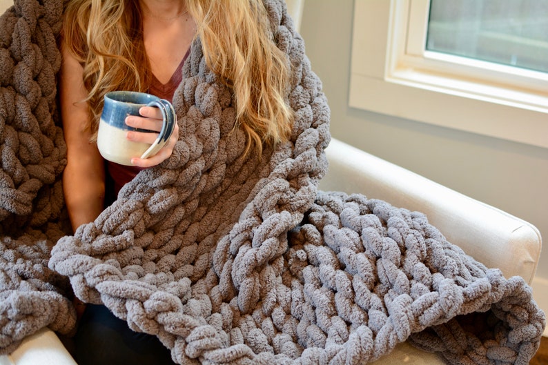 May include: A person is sitting in a chair wrapped in a gray chunky knit blanket. They are holding a blue and white ceramic mug.