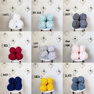 May include: Nine balls of yarn in different colors, including white, spa blue, dark gray, red, light gray, blush pink, navy, gold, and slate. Each ball of yarn is sitting on a small table with a gold geometric design.
