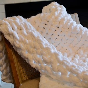 May include: A white chunky knit blanket draped over a wooden chair. The blanket is made of thick, fluffy yarn and has a soft, cozy texture.