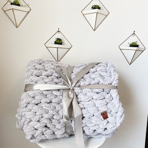May include: A gray chunky knit throw blanket tied with a silver ribbon. The blanket is sitting on a white stool with a wooden base. There are four geometric wall planters with succulents in the background.