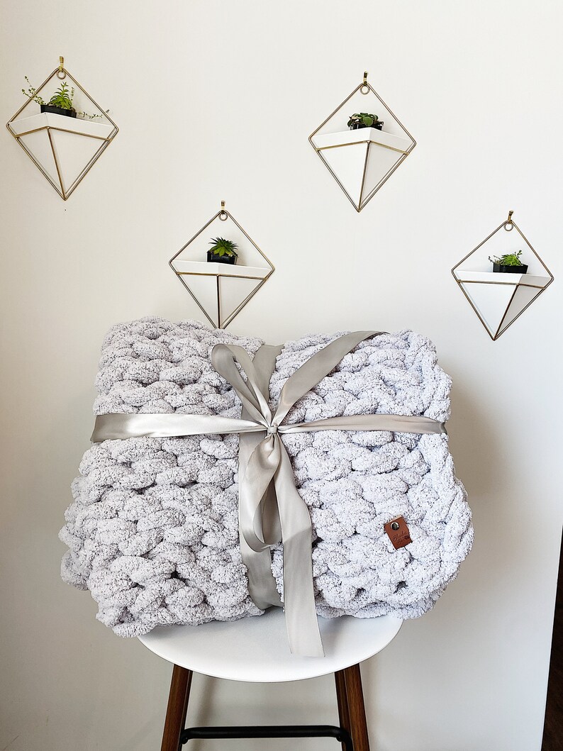 May include: A gray chunky knit throw blanket tied with a silver ribbon. The blanket is sitting on a white stool with a wooden base. There are four geometric wall planters with succulents in the background.