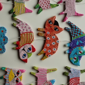 May include: A colorful garland of paper mache cats, each with unique patterns and designs. The cats are strung together with white thread and have pink, orange, blue, green, and yellow accents.
