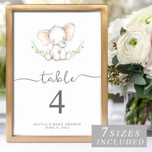 May include: A framed table number featuring a watercolor illustration of a baby elephant. The number 4 is displayed prominently, with the text "table" above it. The frame is gold, and the text "Olivia's Baby Shower June 4, 2022" is included.