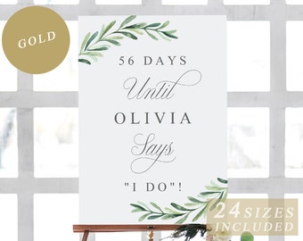 May include: A light blue sign with a green leafy border and the text "56 DAYS Until Olivia Says "I DO"!"  The sign is on a wooden easel.  The text "24 SIZES INCLUDED" is at the bottom of the sign.
