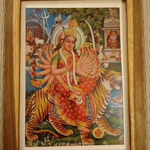 May include: A framed print showing a multi-armed deity on a tiger. The image uses bright colours, with the deity in red and gold clothing, set against a landscape and other figures. The frame is made of light brown wood.