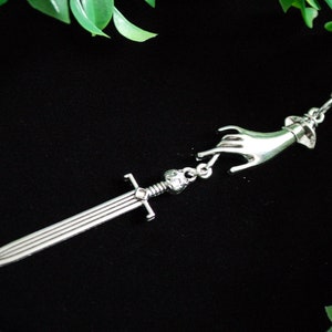 May include: A silver necklace featuring a hand reaching out to hold a sword pendant. The hand and sword are both detailed and have a vintage look.