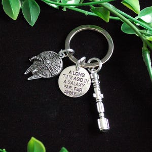May include: Silver-coloured keyring with a Millennium Falcon charm, a lightsaber charm, and a circular charm with the text "A long time ago in a galaxy far, far away." The keyring is on a silver ring.