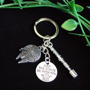 Könnte beinhalten: Silberfarbener Schlüsselanhänger mit einem Millennium Falcon-Anhänger, einem Lichtschwert-Anhänger und einem runden Anhänger mit Yoda und dem Text "MAY THE FORCE BE WITH YOU". Der Schlüsselanhänger befindet sich auf einem schwarzen Hintergrund mit grünem Laub.