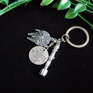 Pode incluir: Chaveiro prateado com um pingente do Millennium Falcon, um pingente de sabre de luz e um pingente circular com o texto "MAY THE FORCE BE WITH YOU" e um gráfico de Yoda. O chaveiro é preso a um anel prateado.