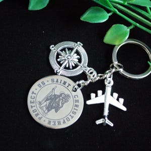 Op de afbeelding: Een zilveren sleutelhanger met drie bedels: een kompas, een ronde Sint-Christoffelmedaillon en een vliegtuig. Op het medaillon staan de woorden "PROTECT US SAINT CHRISTOPHER" rondom. De sleutelhanger ligt op een zwarte ondergrond met groene bladeren.