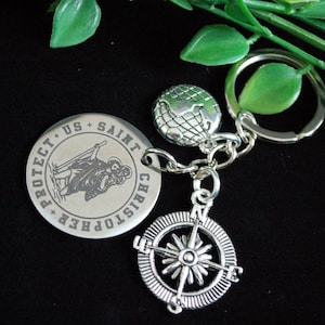 Op de afbeelding: Zilveren sleutelhanger met een kompas, een wereldbol en een ronde bedel met de tekst "PROTECT US SAINT CHRISTOPHER" en een afbeelding van Sint-Christoffel. De sleutelhanger ligt op een zwarte ondergrond met groene accenten.