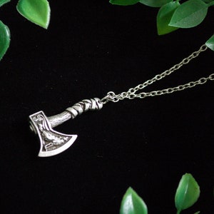 May include: A silver axe pendant necklace with a detailed design on the axe head. The necklace is on a black background with green leaves.