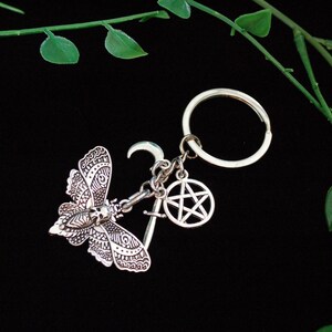 May include: Silver keychain featuring a detailed moth charm, a crescent moon charm, a pentagram charm, and a silver key charm. The charms are attached to a silver key ring.