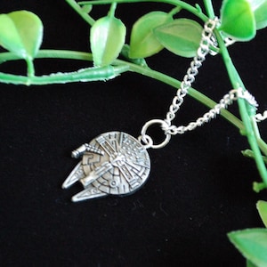 May include: A silver pendant necklace with a Millennium Falcon design. The pendant is suspended from a fine chain. The necklace is set against a black background with green foliage.