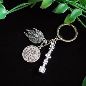 May include: A silver-colored keychain featuring three charms: a Millennium Falcon, a lightsaber, and a circular charm with the text "THAT'S NO MOON". The keychain is attached to a silver ring. The background is black with green leaves.