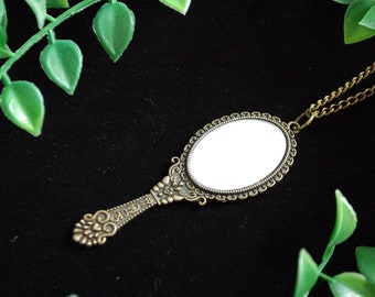 May include: A gold-toned ornate hand mirror necklace with a chain. The mirror is oval shaped and has a decorative frame. The necklace is on a black background.