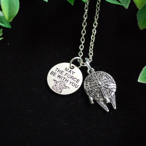May include: Silver-toned necklace featuring two pendants: a round charm with Yoda and the text "MAY THE FORCE BE WITH YOU," and a Millennium Falcon pendant. The necklace hangs against a black background, with green foliage in the corners.