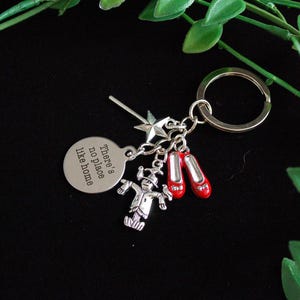 May include: Silver-toned keychain with charms inspired by The Wizard of Oz. Charms include a pair of red shoes, a scarecrow, a star, and a circular charm with the text "There's no place like home."