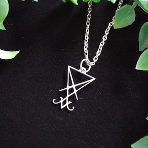 Op de afbeelding: Een zilveren ketting met een hanger met een geometrisch symbool, mogelijk een sigil of een symbool van een godheid. De hanger heeft de vorm van een driehoek met lijnen en krommen erin. De ketting is gefotografeerd tegen een zwarte achtergrond met groene bladeren.