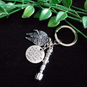 May include: Silver keychain featuring a Millennium Falcon charm, a lightsaber charm, and a circular charm with the text "A long time ago in a galaxy far, far away...". The keychain is on a black surface with green leaves.