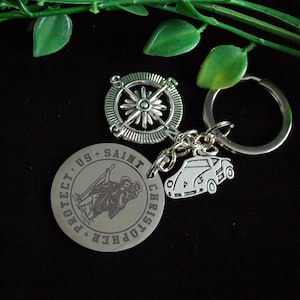 Op de afbeelding: Een verzameling zilverkleurige sleutelhangers en bedels. Bevat een kompasontwerp, een auto-bedel en een ronde bedel met de tekst "PROTECT US SAINT CHRISTOPHER". De items zitten aan een sleutelring.