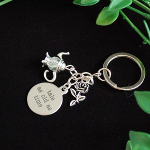 Pode incluir: Chaveiro prateado com pingentes, incluindo uma chaleira, uma rosa e uma etiqueta redonda com o texto "tale as old as time". O chaveiro está em um fundo preto com folhagem verde.