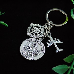 Op de afbeelding: Zilveren sleutelhanger met een kompas, vliegtuig en een ronde hanger met de woorden "Sint Christoffel Bescherm ons".