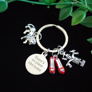 May include: Silver-toned keychain with charms. Charms include a lion, a scarecrow, and a pair of red shoes. A round charm is engraved with the phrase "There's no place like home". The keychain is on a black background.
