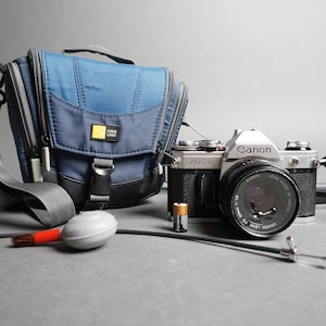 May include: A silver Canon AE-1 camera with a lens attached, a blue camera bag with a strap, and a grey air blower. The camera has a black strap attached to it.