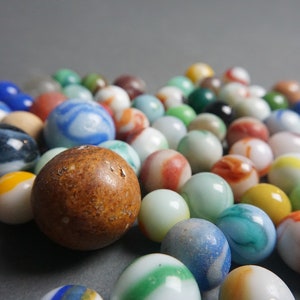 May include: A collection of colorful glass marbles in various patterns and colors, including blue, green, red, white, and brown. The marbles are arranged in a loose pile on a gray surface.