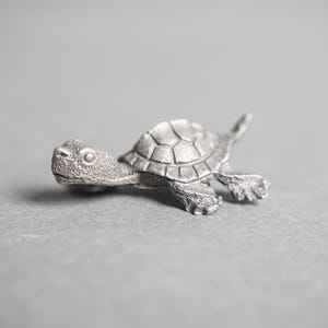 May include: A silver turtle figurine with a detailed shell and head. The turtle is in a swimming position with its head and legs extended.